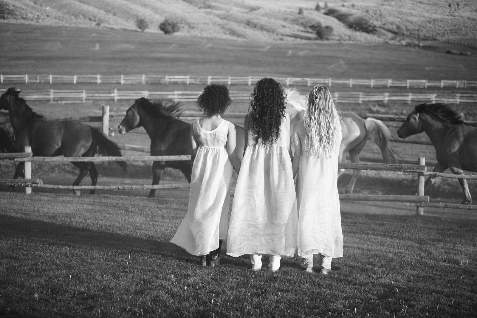Three women in white dresses watching horses galloping past