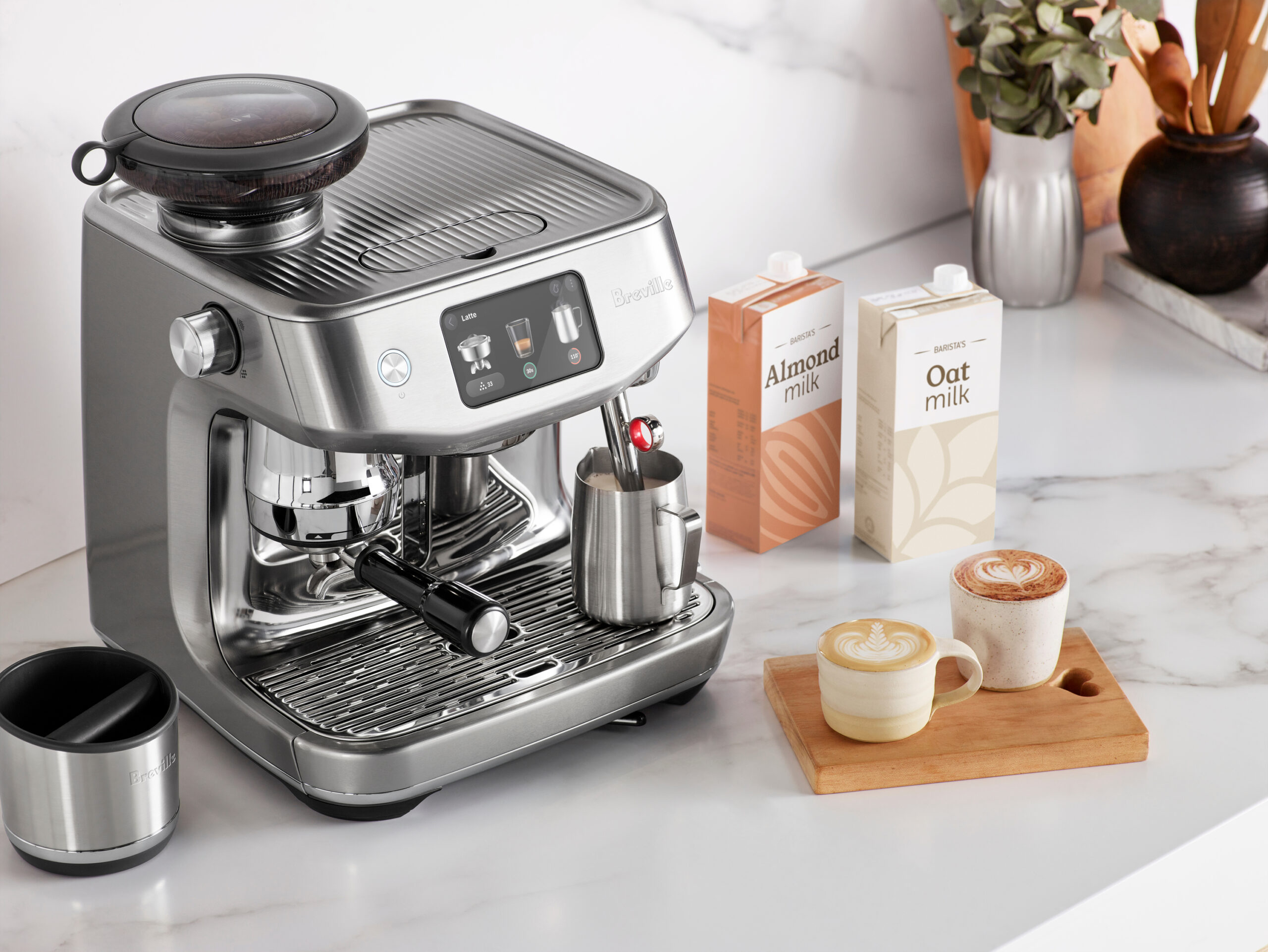 An espresso maker on a counter with two cups of prepared coffee and containers of oat and almond milk.