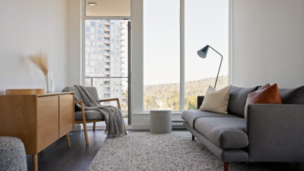 The interior of an apartment. A couch, credenza, chair, and lamp in front of a window.