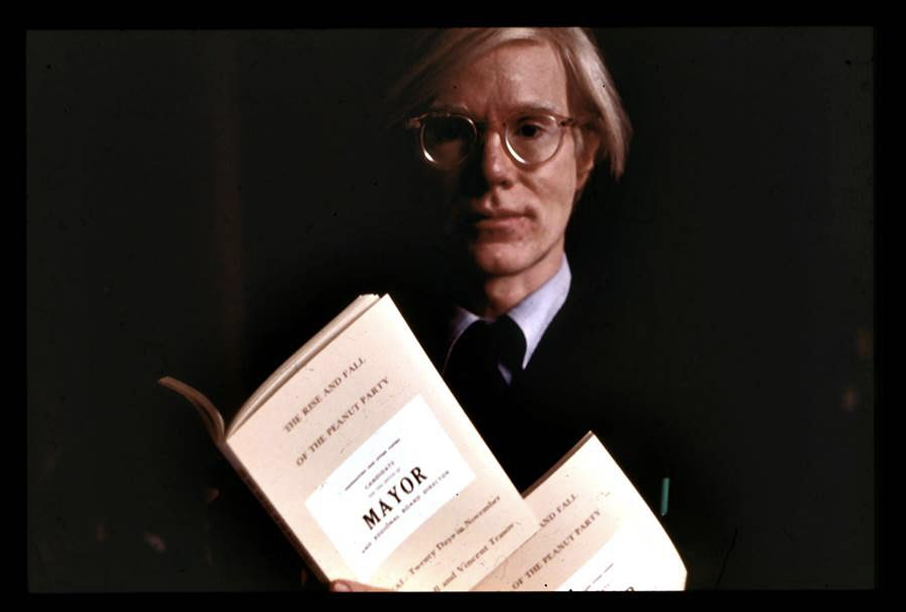 Andy Warhol holding a booklet titled The Rise and Fall of the Peanut Party.