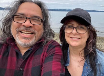 A couple looks into the camera, smiling, with a BC beach in the background.