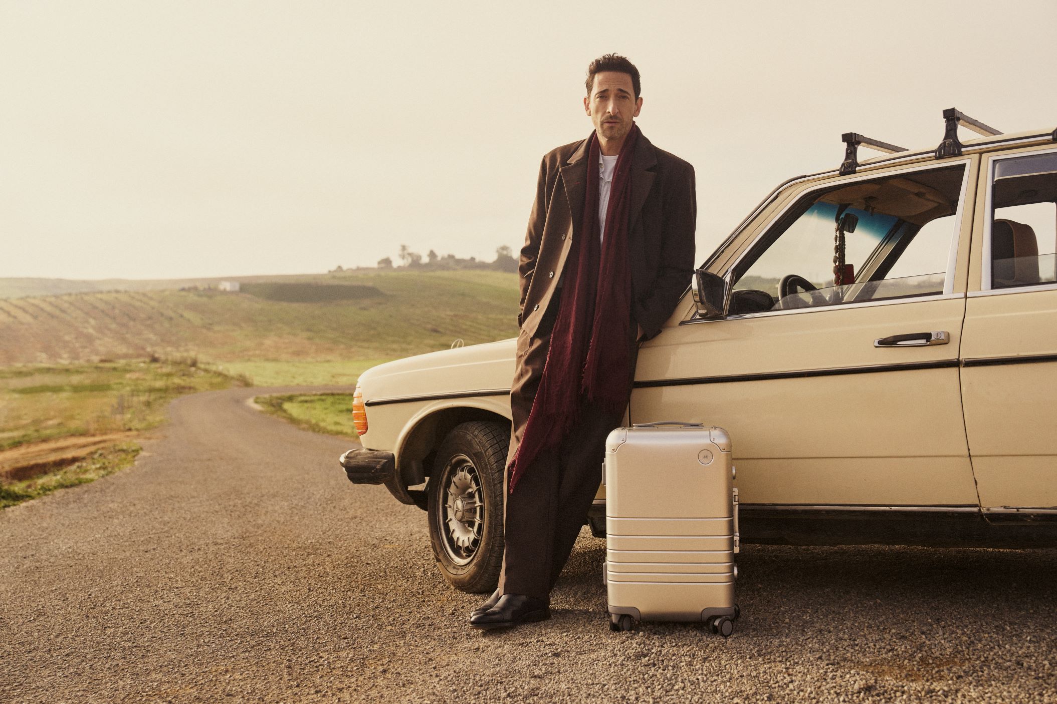 A man leans against a car with a piece of Monos luggage - April wishlist