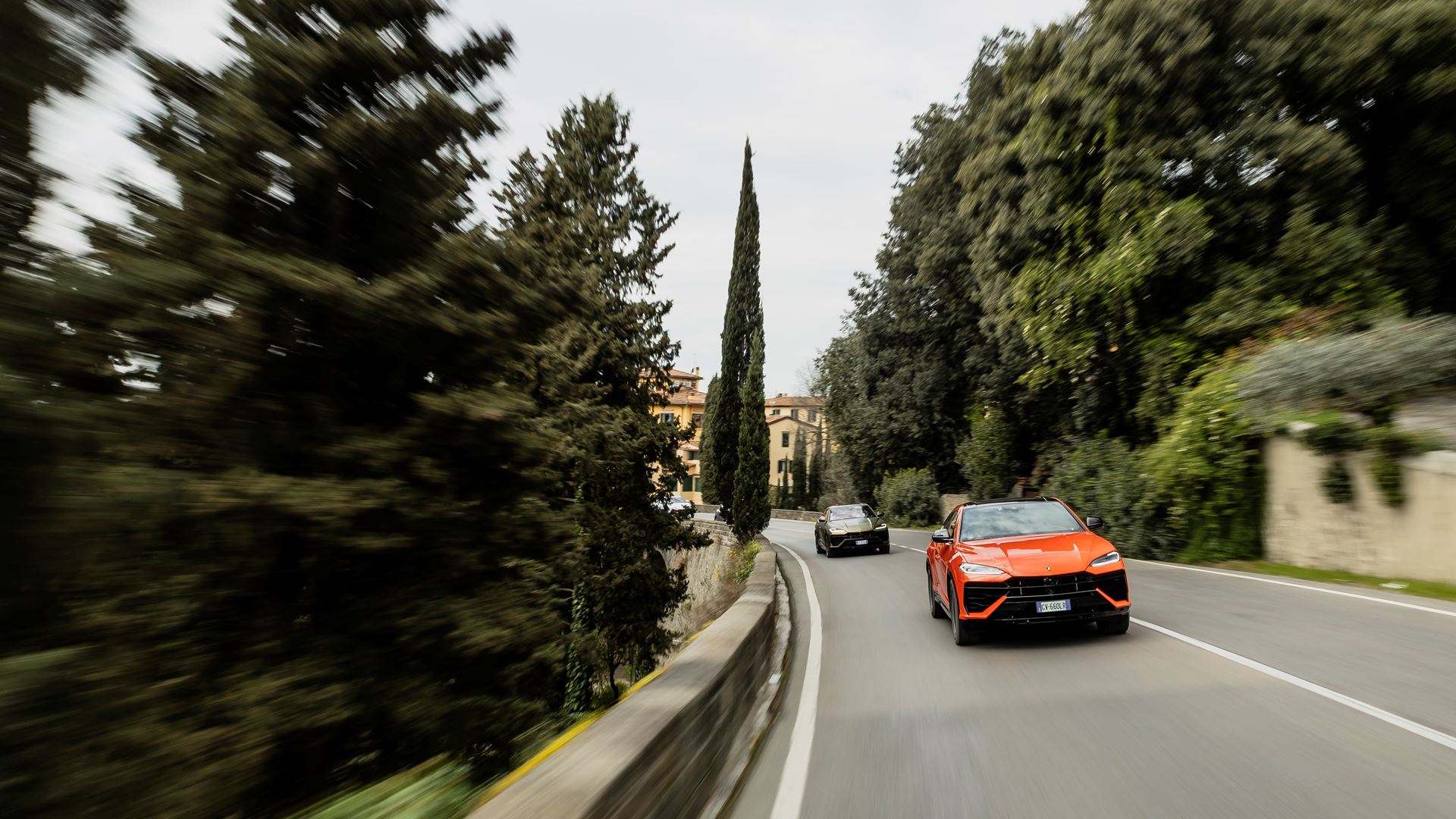 An orange Lamborghini Urus SE speeds down a road ahead of a black car.