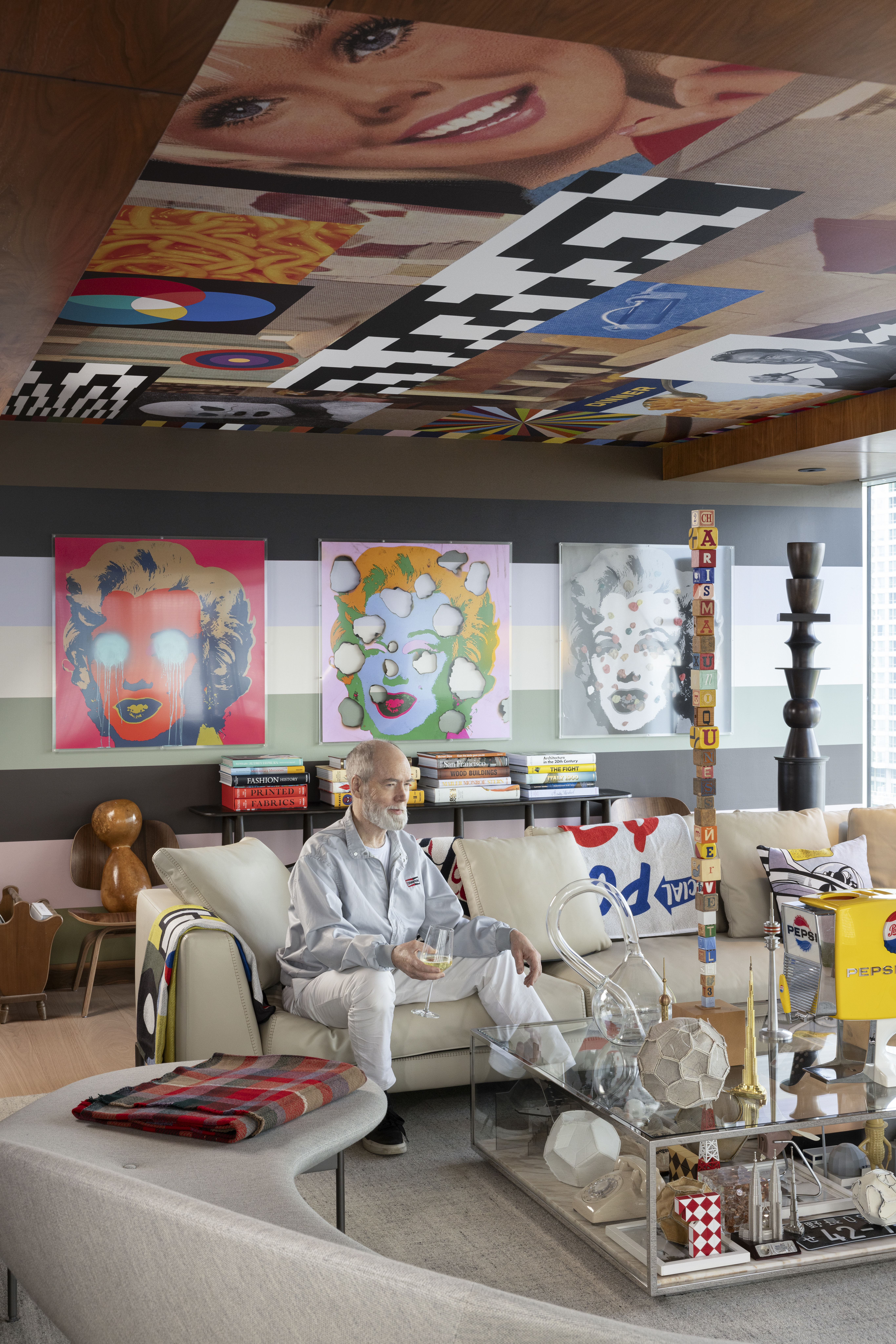Douglas Coupland sits on a couch, holding a glass, in Suite X, surrounded by pop art.