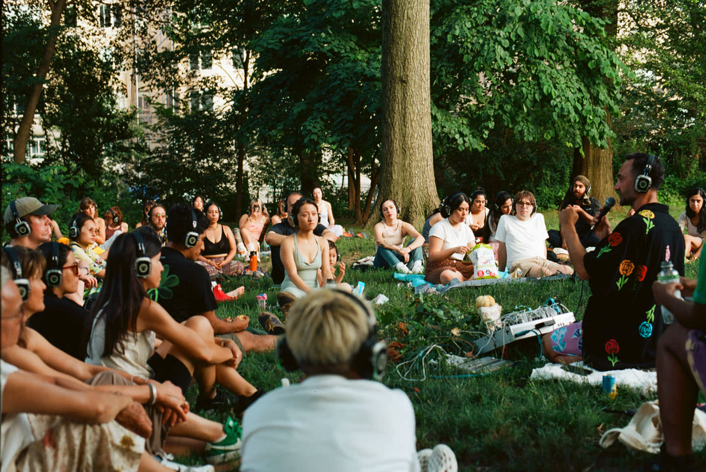 A group of people sit outdoors wearing headphones, positioned around Tarun Nayar (Modern Biology) as he plays music on a piece of machinery.