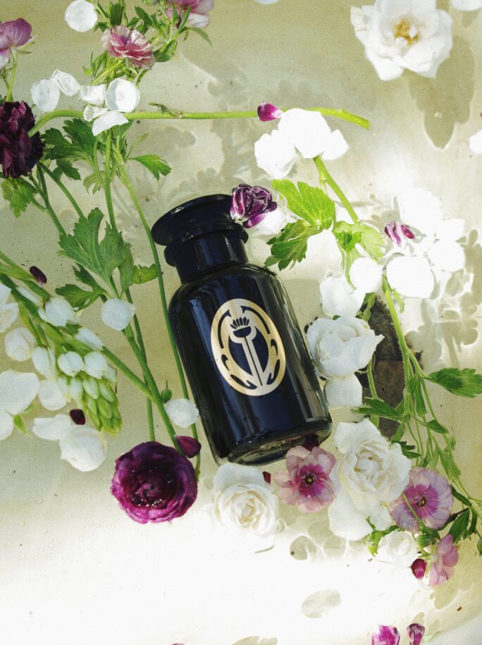A black glass bottle of Sangre de Fruta bath salt in a tub full of flowers.