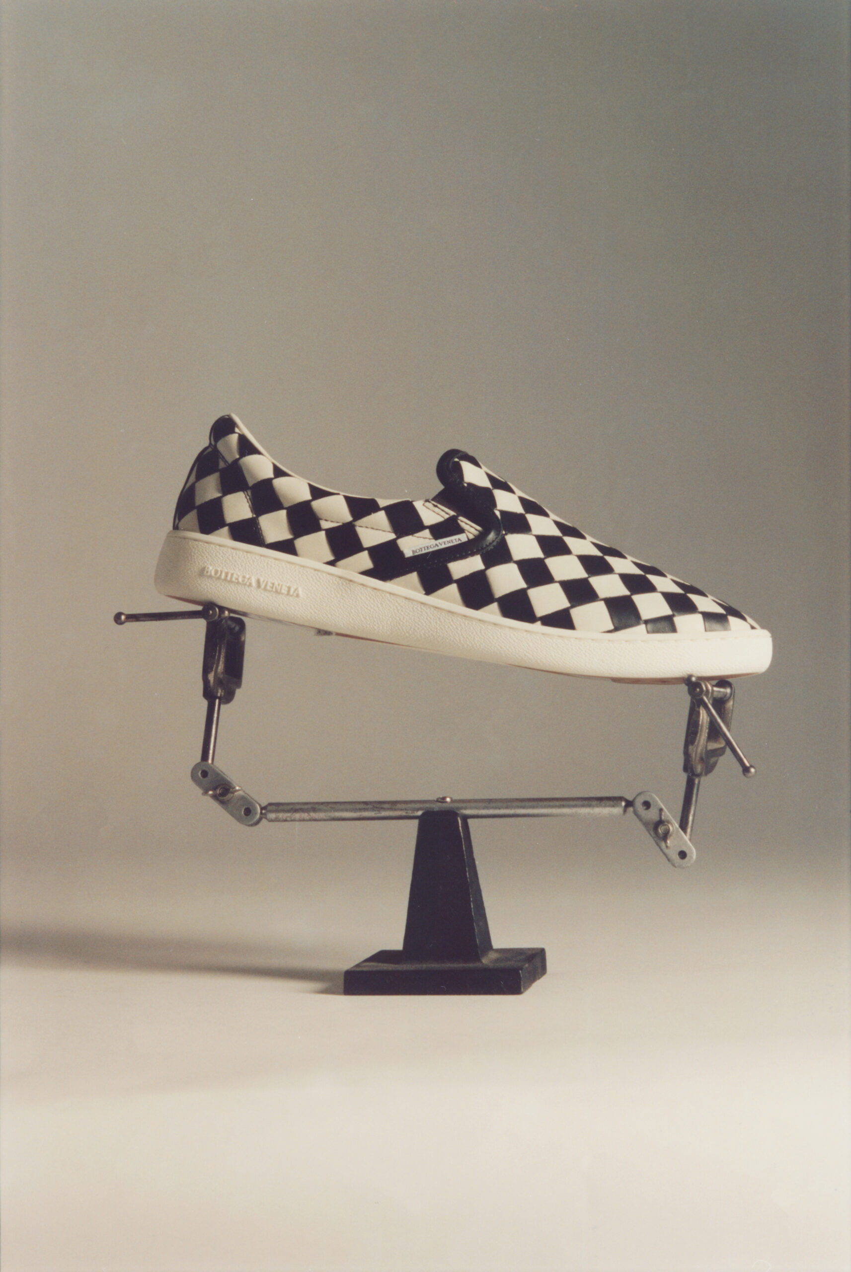 A men's shoe, slip on with black and white checkers, help up on display in a metal tool of some kind.