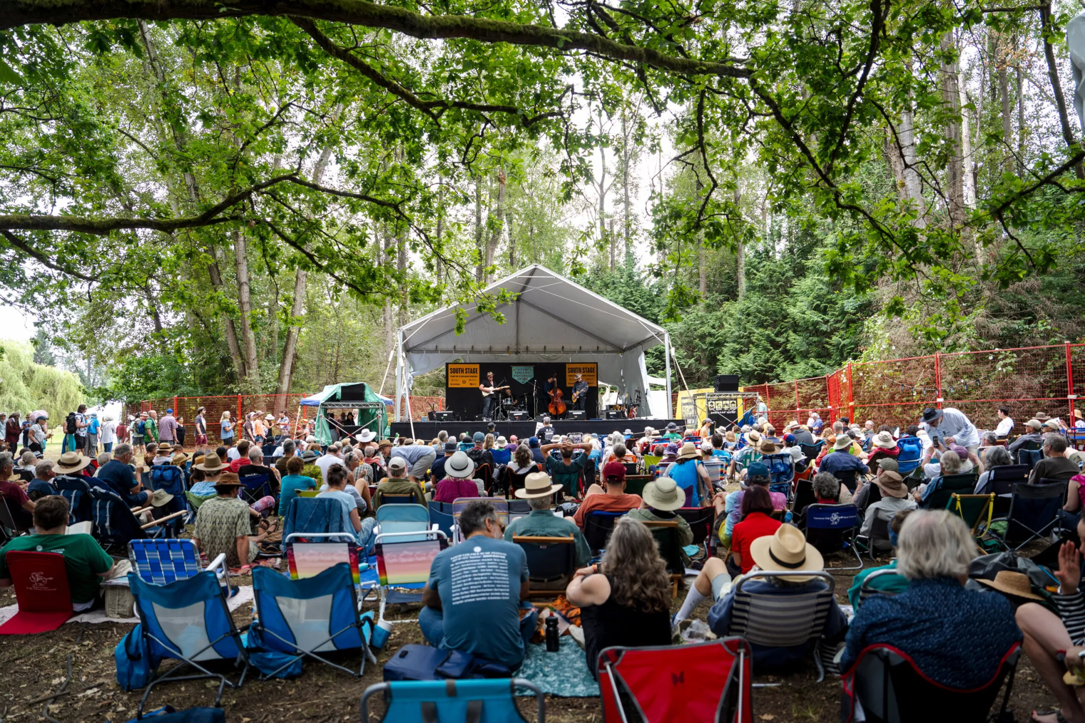 People in lawn chairs watch a tented stage surrounded by trees.