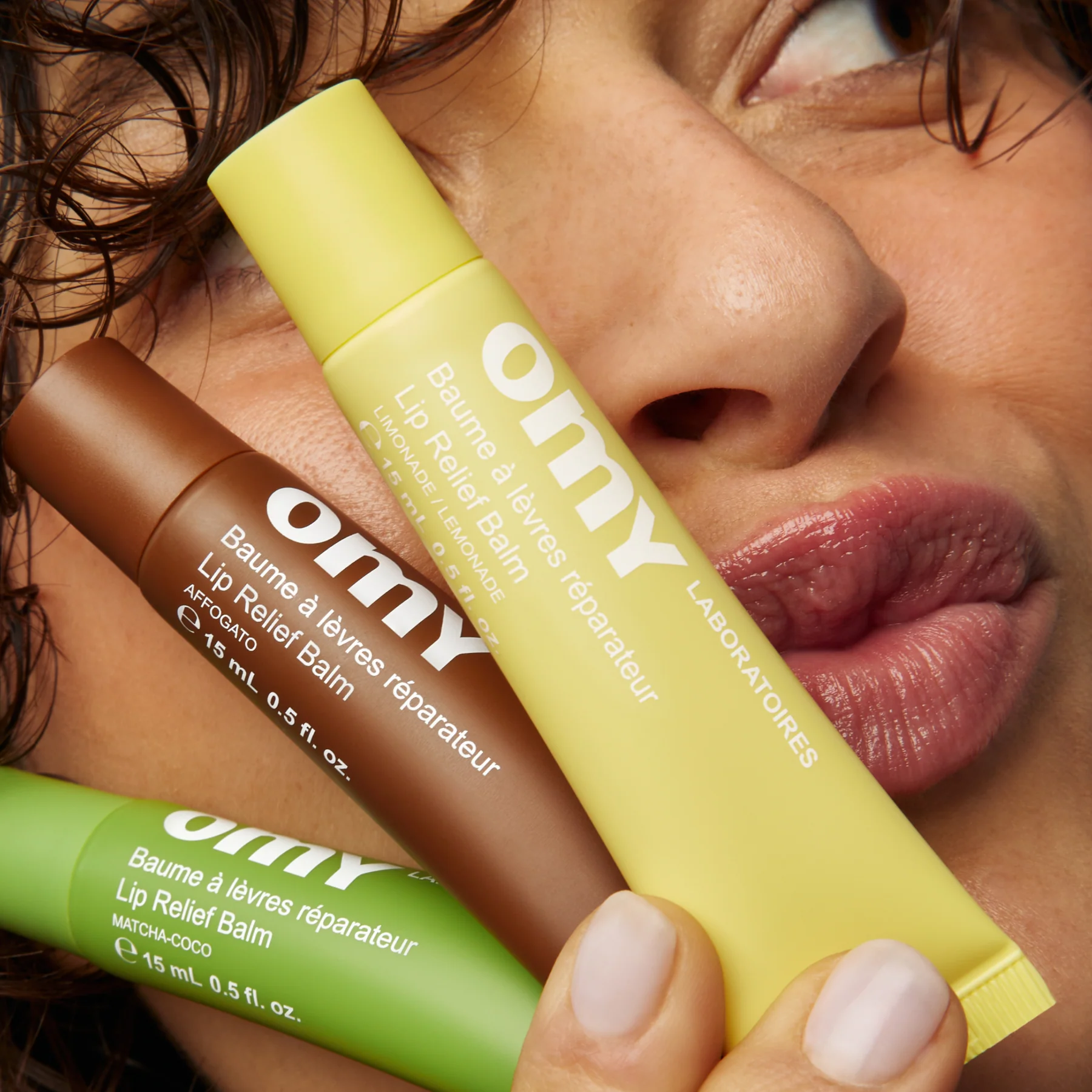 A closeup of a woman's face, holding up three tubes of Omy products