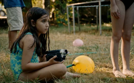 A girl sits in the grass in a backyard holding a video camera.