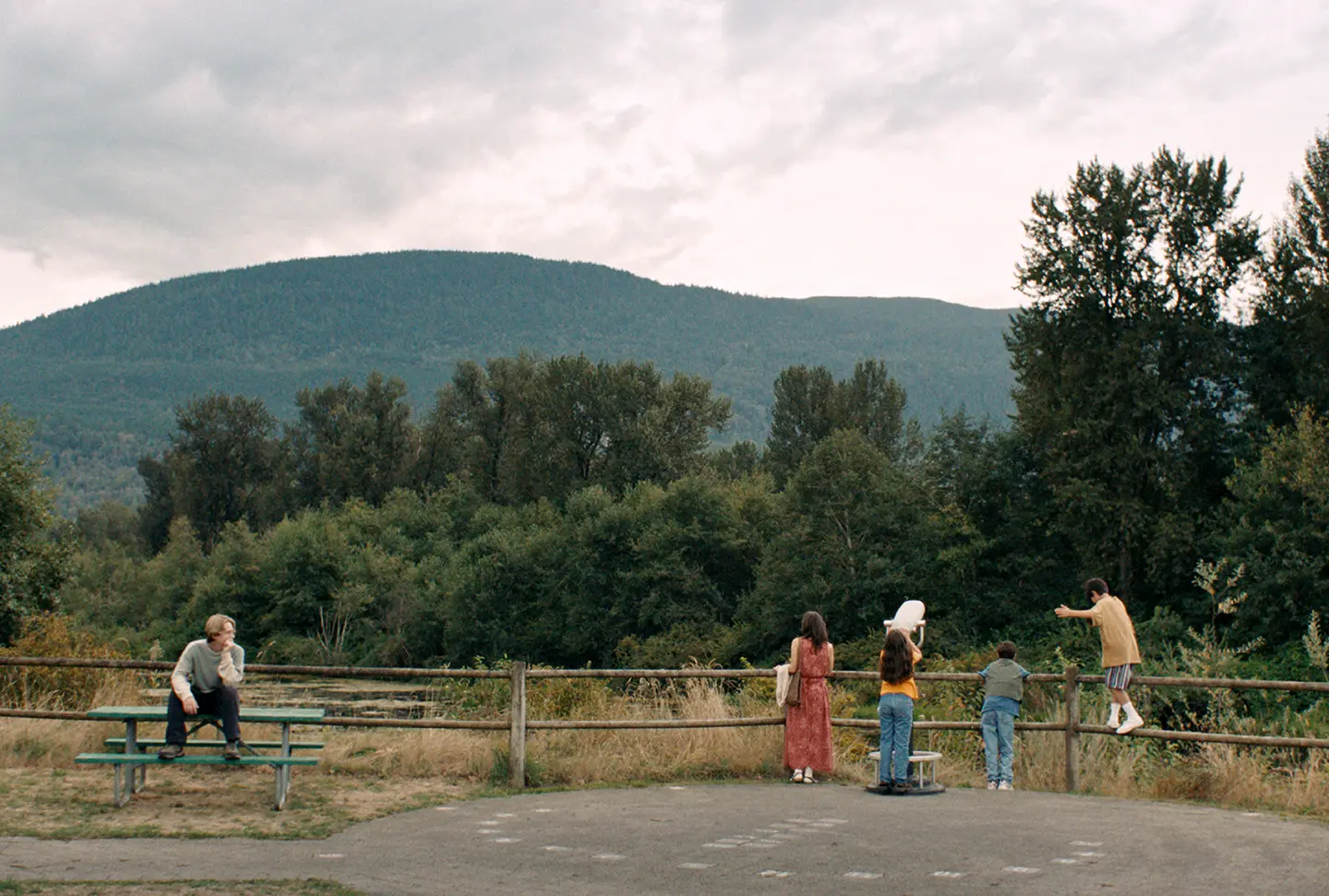 A young man sits on a picnic table while four other people stand away from him at a telescope in a still from Blue Heron.