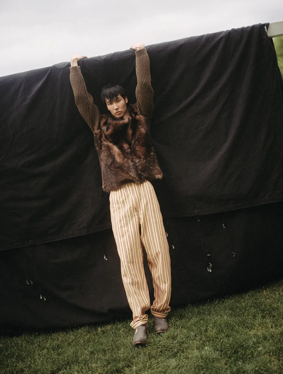 A man leans against a tarp, his arms up as if hanging from it, while looking into the camera.