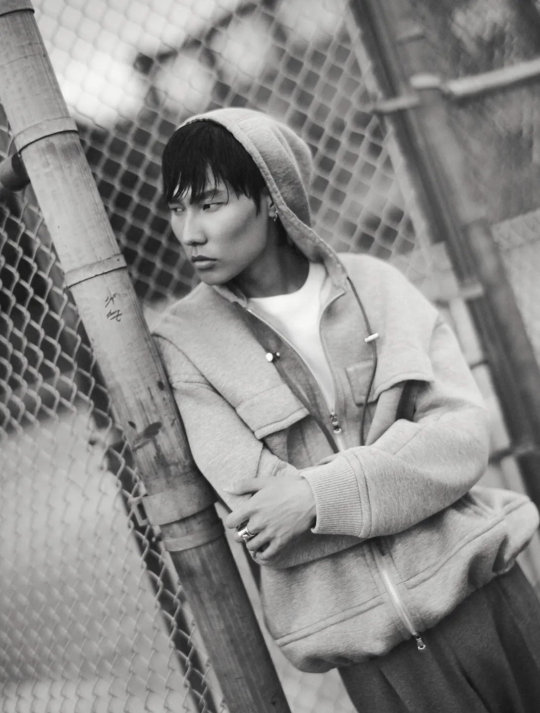 A black and white fashion shoot photo of a man in a hoodie leaning against a chain link fence.