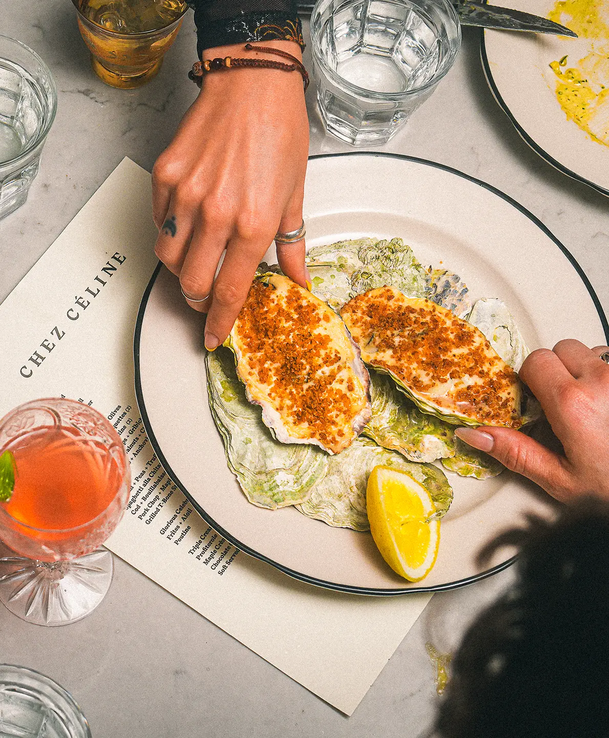 Two hands reach for baked oysters from a plate atop a Chez Céline menu.