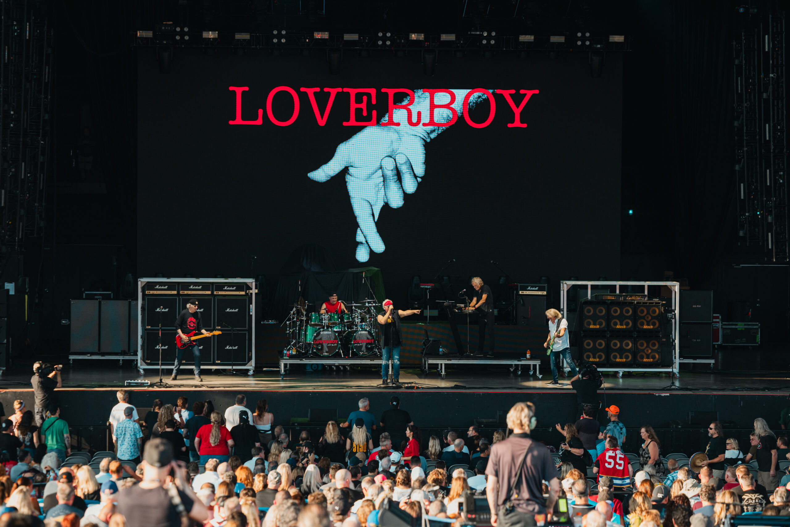 The band Loverboy performs at an outdoor show in front of a large banner labelled "Loverboy."