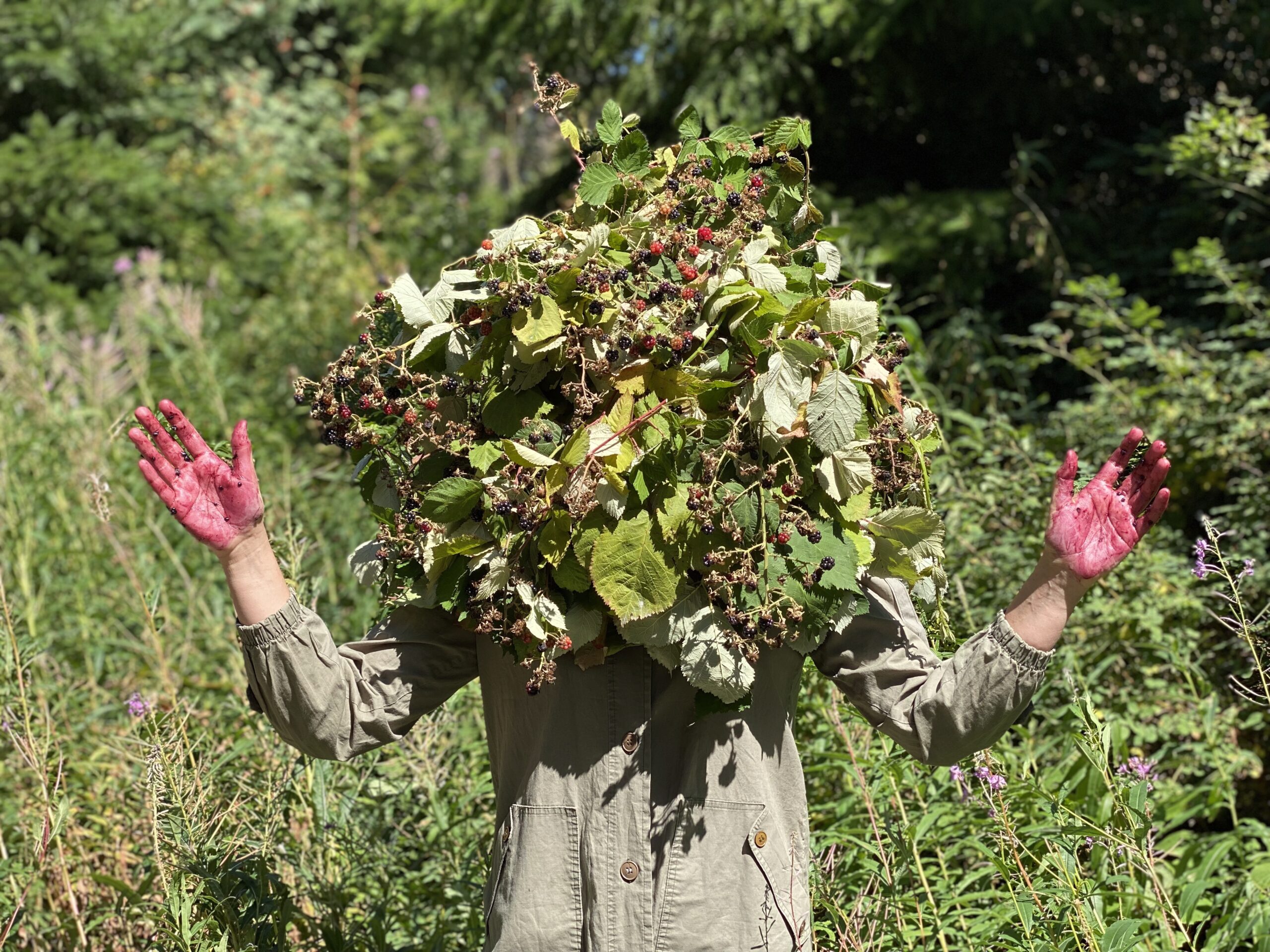 A woman in a beige jumpsuit wears a blackberry bush on her head. Her outstretched hands are covered in berry juicer.