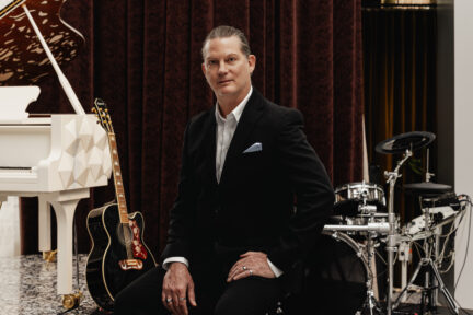 A man in a suit (Rob Eller) sits at a piano, surrounded by intruments.