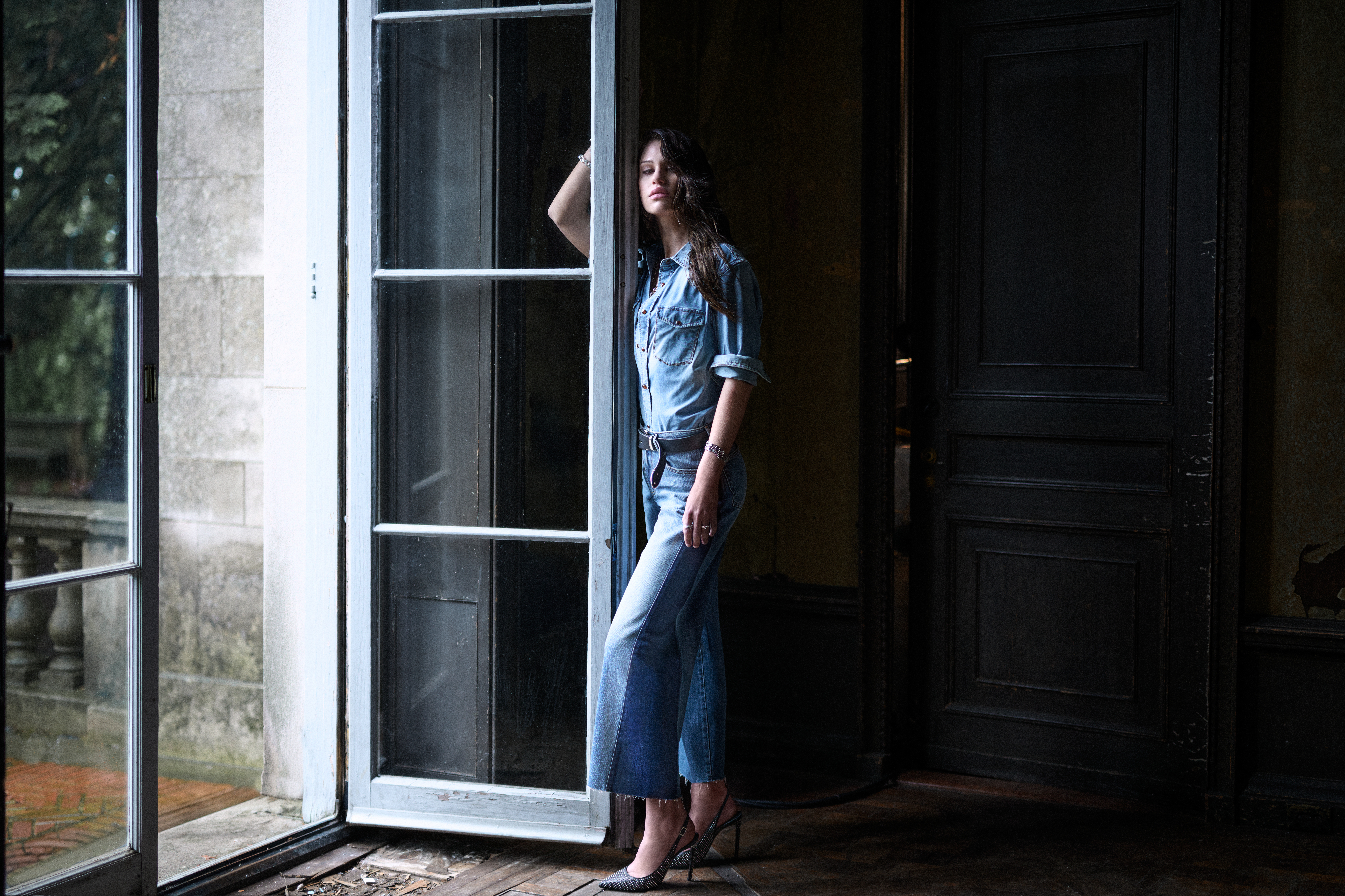 A woman leans against an open door wearing denim pants and shirt.
