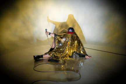 A musician masked in a glittery shawl poses on stage with a microphone - Jeff Cancade as the Golden Age of Wrestling