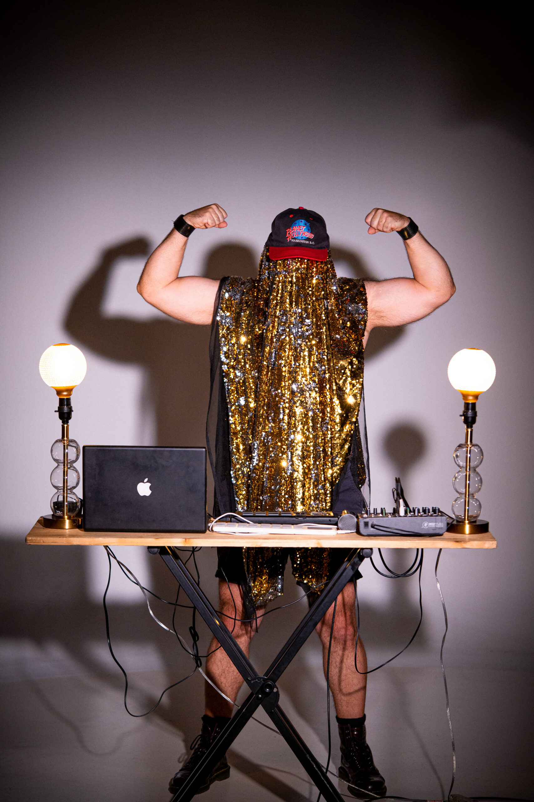 Golden Age of Wrestling stands at a DJ table, holding their arms up with flexed biceps.