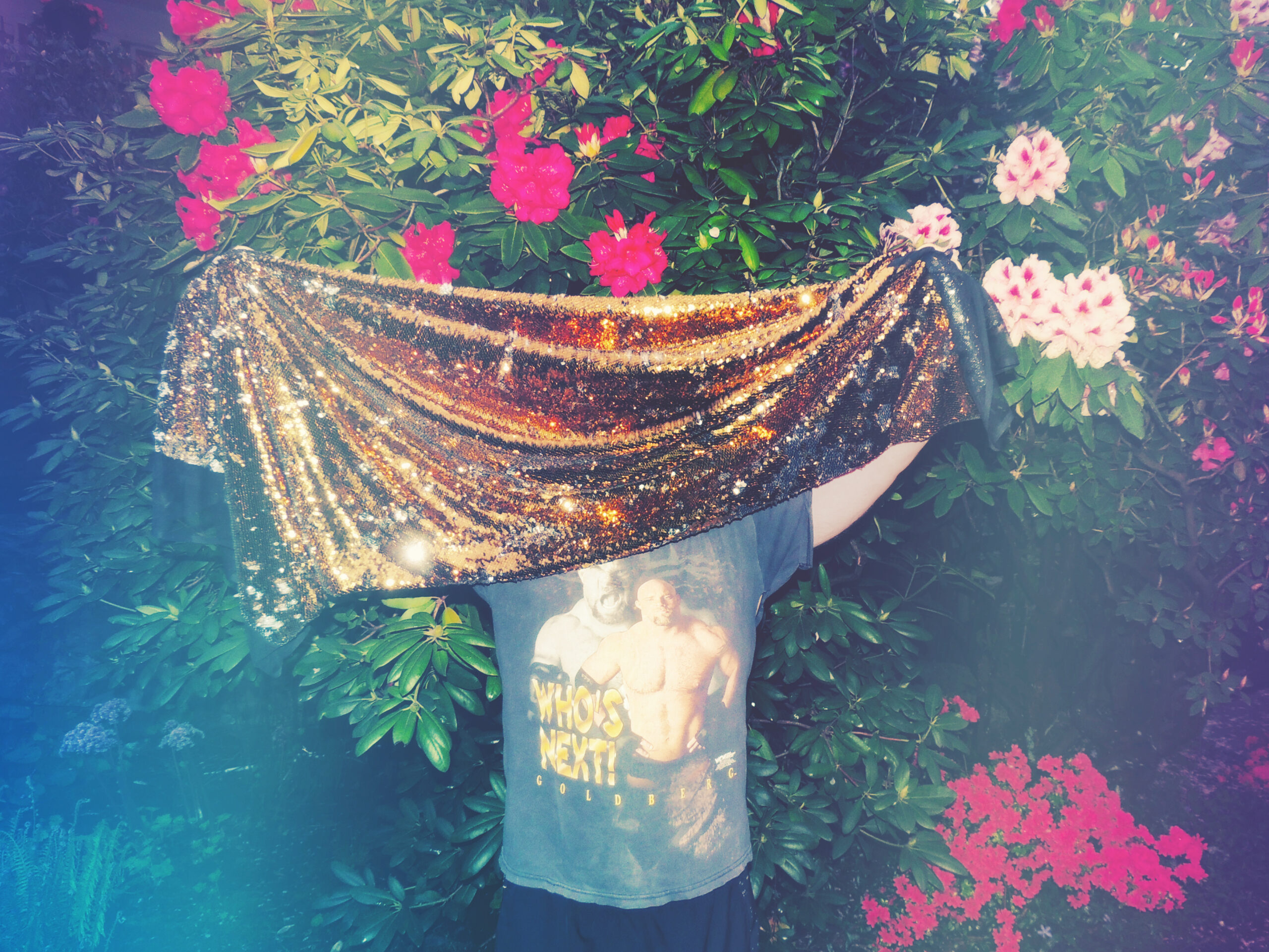 the Golden Age of Wrestling holds up a glittery shawl in front of their face in a flower garden.